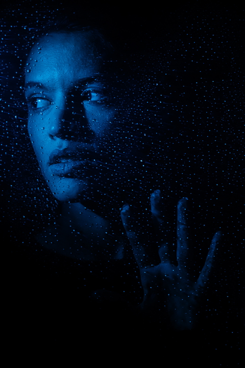 A woman's face and hand appear behind a wet glass surface, creating a moody and intimate atmosphere.