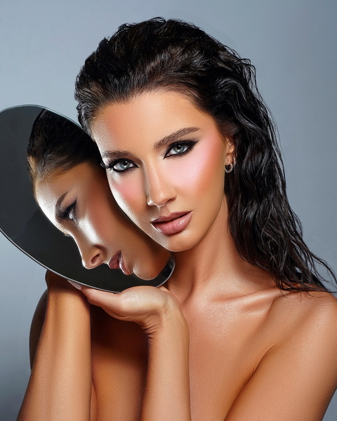 A beautiful woman poses with a mirror, showcasing her elegant makeup and flawless skin in a glamour shot.