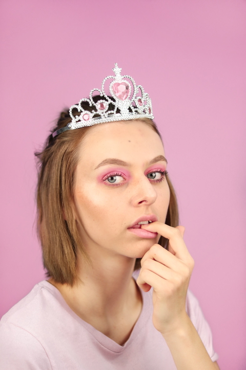 A young woman with pink makeup poses with a playful expression, wearing a princess crown against a pink backdrop.