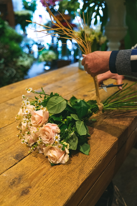 A close-up of a delicate bouquet of peach roses and chamomile being arranged at a florist shop on a wooden table.