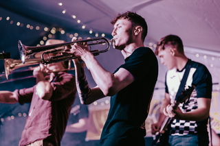 Three musicians perform on stage, playing a trumpet, trombone, and guitar during a lively indoor concert.