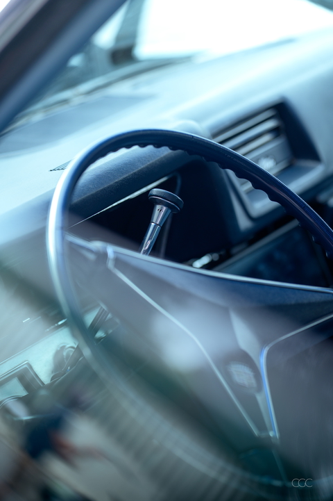 A classic car's blue steering wheel and silver gear stick are captured in a close-up, vintage-style shot.