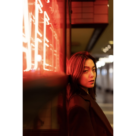 A woman leans against a vibrant red neon light in an urban setting, looking intensely at the camera.