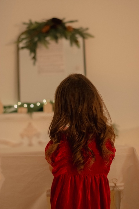 A little girl in a red dress looks at the mantel, decorated for Christmas.