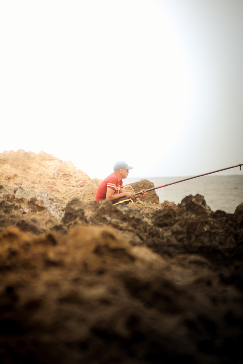A man wearing a red shirt fishes from a rocky coast, looking out at the ocean.