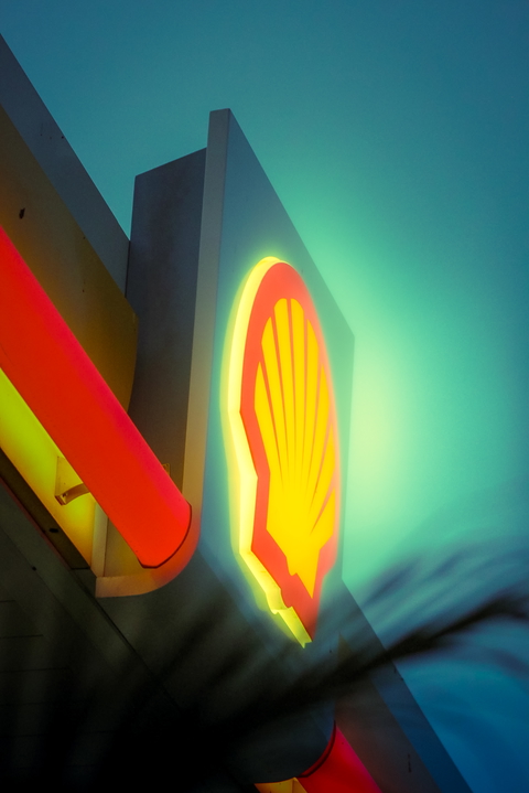 A brightly lit Shell gas station logo stands out against a tranquil blue and green sky.