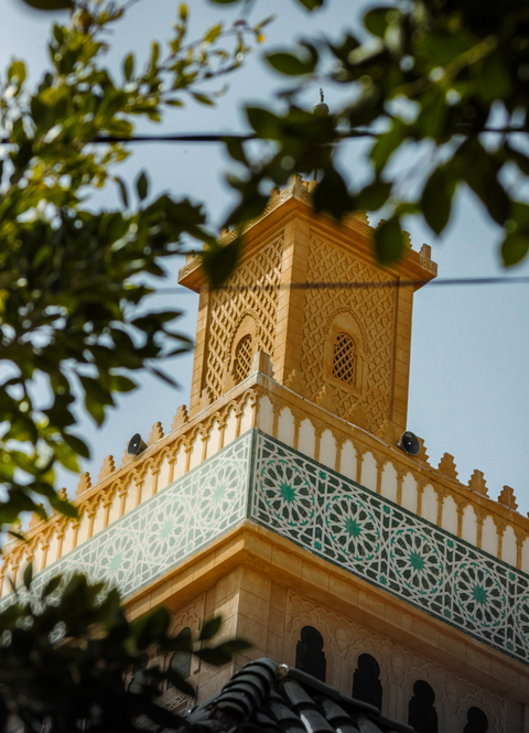 A low-angle view of a beautifully decorated mosque minaret against a clear sky, framed by tree branches.