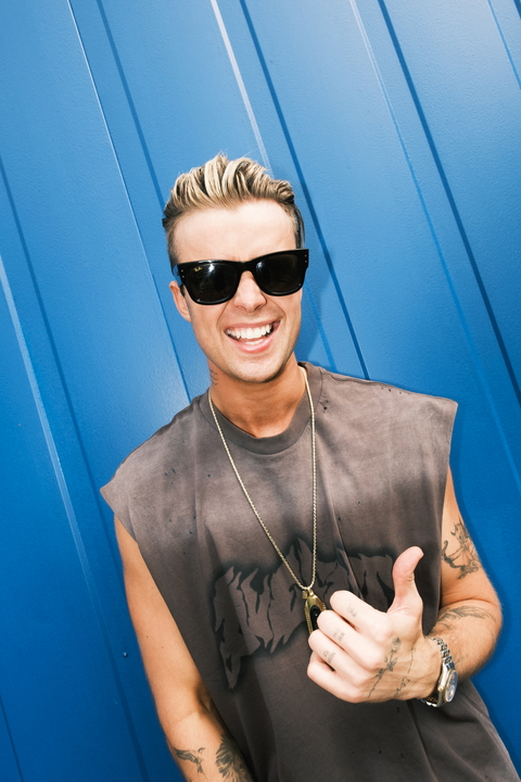A man with sunglasses smiles and gives a thumbs up in front of a blue wall.