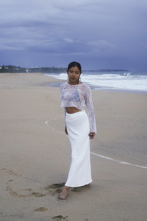A woman in a white skirt and top stands on a sandy beach with the ocean in the background.
