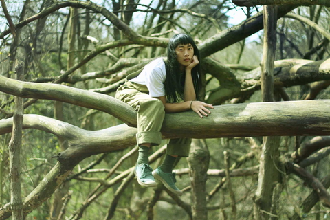 A person wearing olive green overalls sits thoughtfully on a tree branch in the woods.