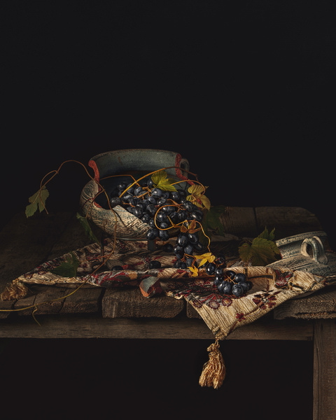 A still life composition with grapes in a pottery bowl on a wooden table with an ornate tablecloth.