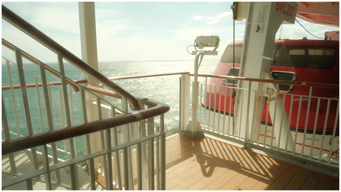 An ocean view from the deck of a ship. Features a lifeboat, metal railings, and the sea. 